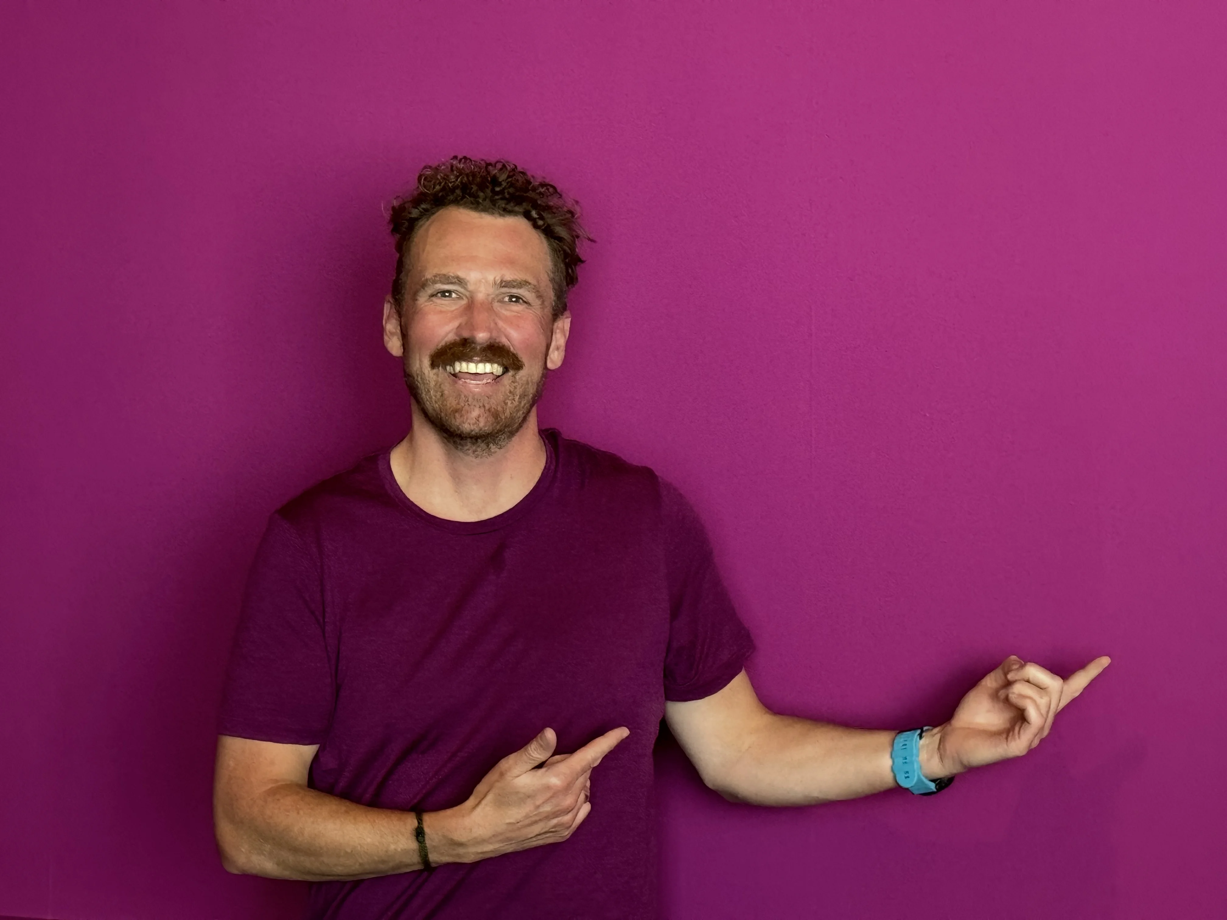Trent smiling and pointing to the side against a magenta wall